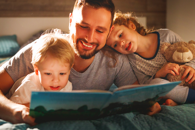 Vediamo una bambina ed un bambino leggere un libro insieme al loro padre sul letto.