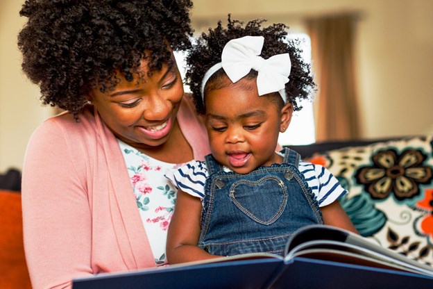 Vediamo una mamma sorridente leggere un libro alla propria figlia.